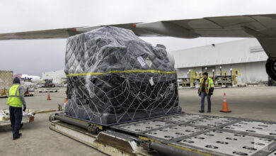 A large pallet, shrink-wrapped in black plastic rests on the tarmac under the wing of a plane.