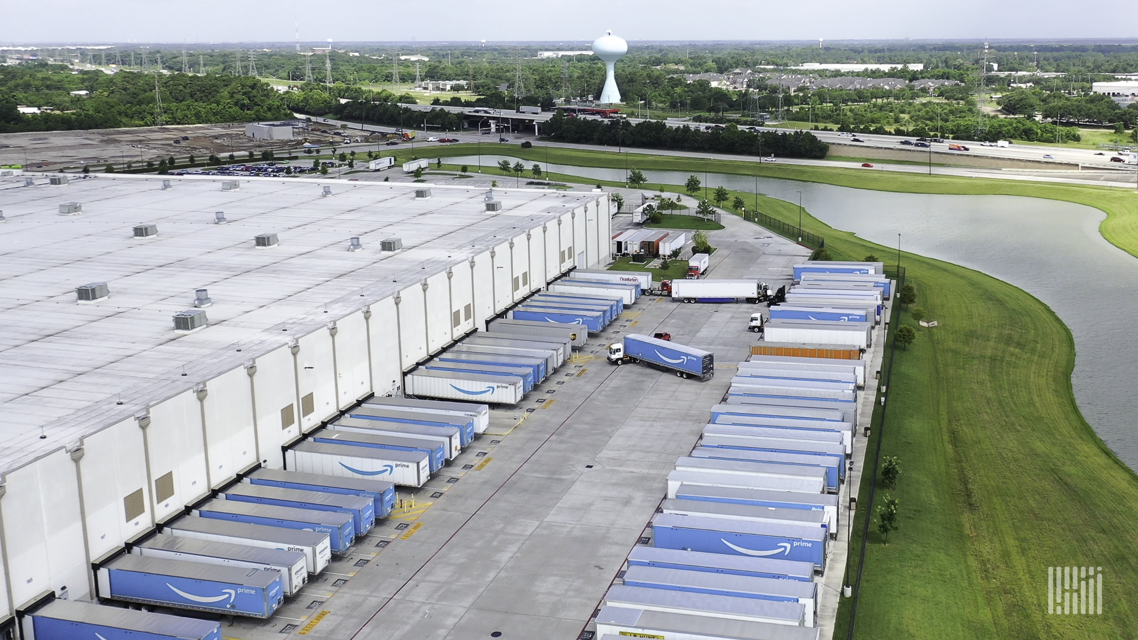 An Amazon warehouse with semi trailers parked in the lot sits in front of a green background.