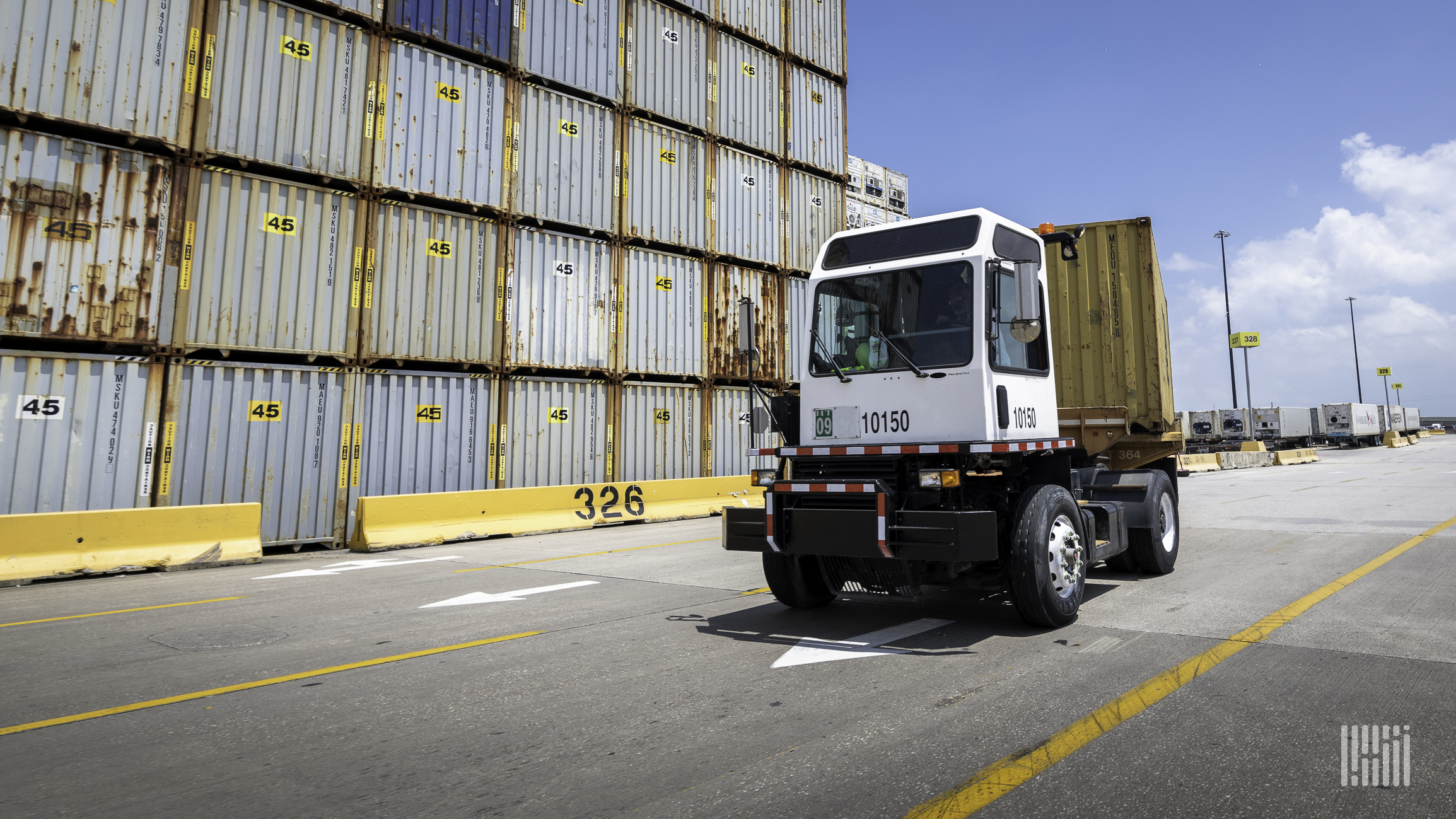 A terminal tractor moves a container at a port with shipping containers in the background.