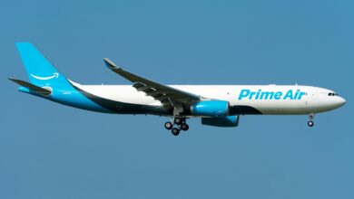 A light-blue tailed Amazon Prime cargo jet approaches airport with wheels down, side view.