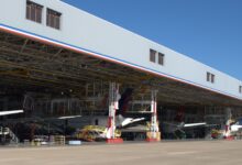View of giant aircraft hangar with several aircraft inside, viewed from outside the entrance on a sunny day.