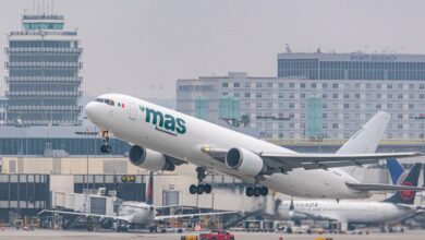 A white "mas" cargo jet takes off from Los Angeles airport with buildings in the background on a gray day.