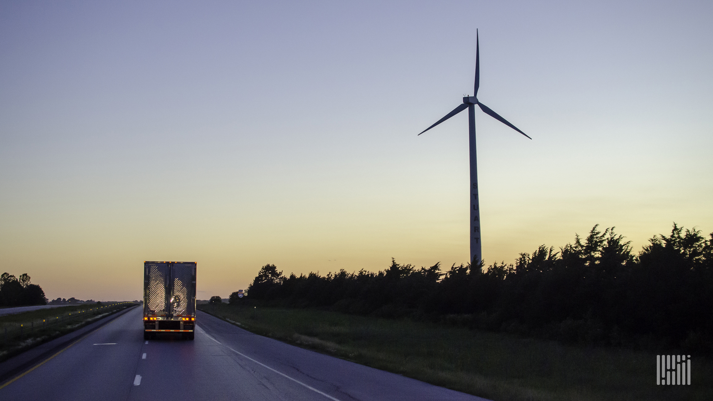 A semi-truck drives down the road with a sunset and wind turbine in the landscape.